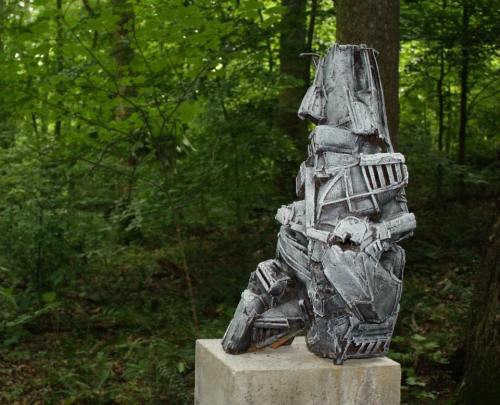 A metallic sculpture resembling a seated figure, surrounded by lush green trees in a forest setting.
