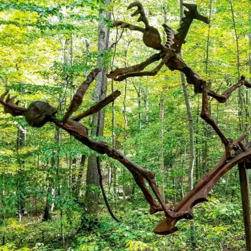 Two whimsical metal sculptures of figures in a forest, one appearing to chase the other among green trees.