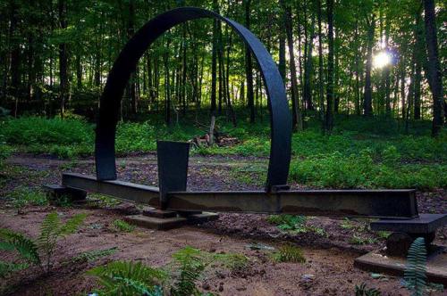 A large, circular metal sculpture stands in a lush green forest, with sunlight filtering through the trees.