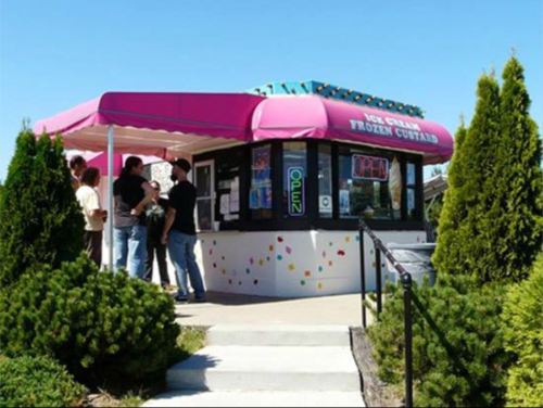 A colorful ice cream stand with a pink awning, people lined up outside, surrounded by green shrubs.
