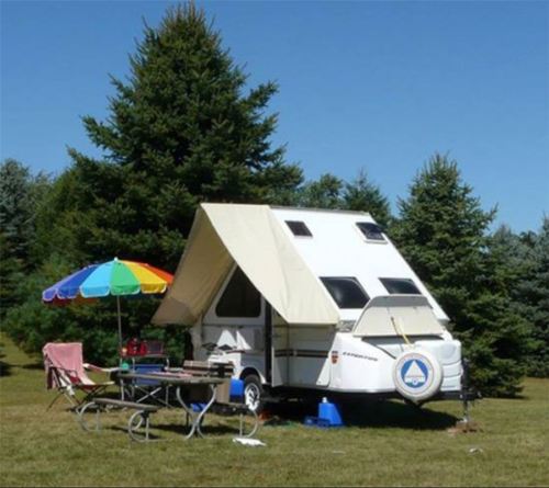 A cozy camper trailer set up in a grassy area, surrounded by tall trees, with a colorful umbrella and picnic table nearby.