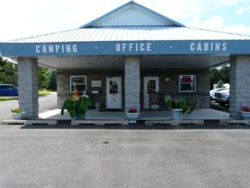 A building with a sign reading "Camping Office Cabins," featuring a gray roof and stone pillars, surrounded by greenery.