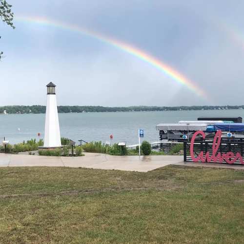 A scenic view of a lake with a lighthouse, boats, and a rainbow in the sky, alongside a sign that reads "Culvers."