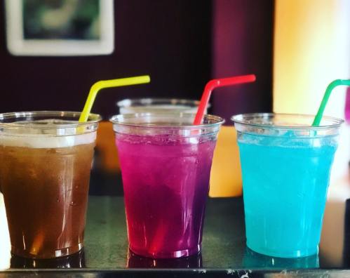 Three colorful drinks in clear cups with straws: brown, purple, and blue, set against a blurred background.
