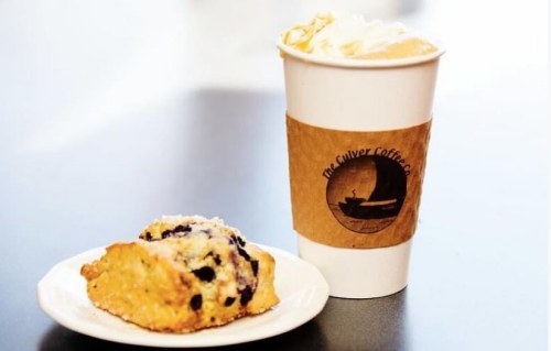 A scone with blueberries on a plate next to a coffee cup with a logo, set on a dark table.