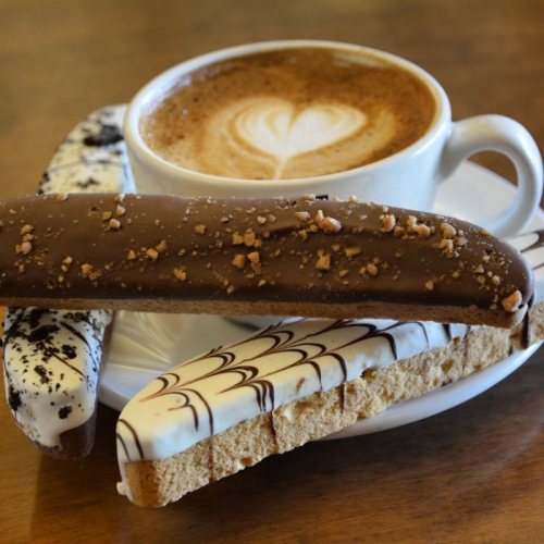 A cup of coffee with a heart design, accompanied by three chocolate-covered biscotti on a saucer.