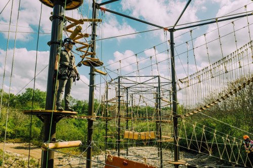 A person in safety gear stands on a platform in a high ropes course, surrounded by trees and blue skies.