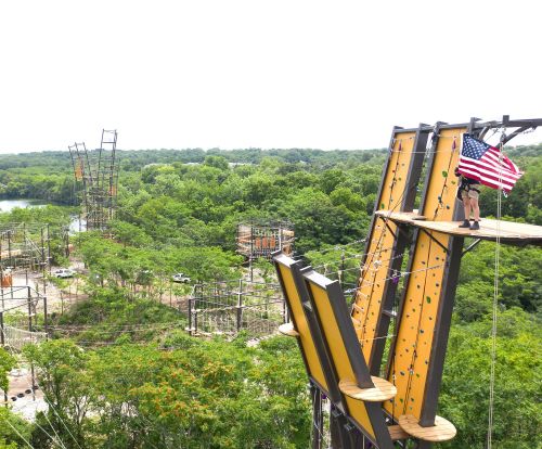 A person stands on a high ropes course platform, holding an American flag, surrounded by trees and adventure structures.