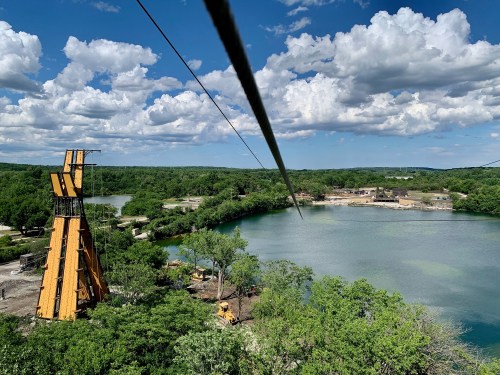 A scenic view of a lake surrounded by greenery, with a zip line tower and clouds in the blue sky above.