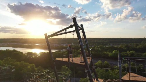 A person stands on a platform at a high ropes course, with a sunset and lush landscape in the background.