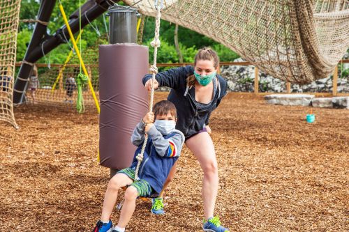 A woman helps a child swing on a rope at a playground, both wearing masks, surrounded by wood chips and climbing structures.