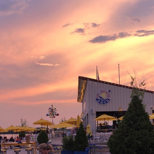 A vibrant sunset over a lakeside venue with yellow umbrellas and a colorful wind spinner.