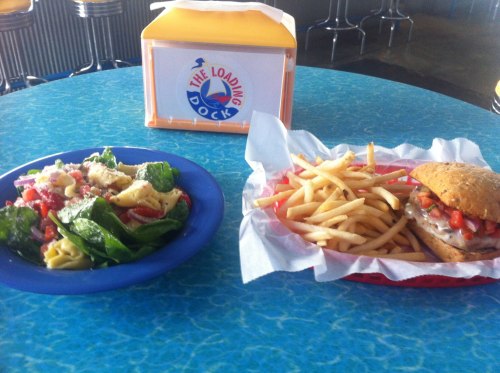 A colorful salad with pasta and spinach next to a sandwich with fries on a blue table, with a drink container in the background.