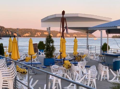 A lakeside venue with yellow umbrellas, white chairs, and a tall wooden sculpture against a sunset backdrop.