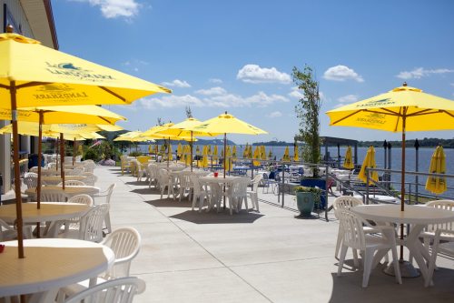 Outdoor dining area by the water, featuring white tables and yellow umbrellas under a clear blue sky.