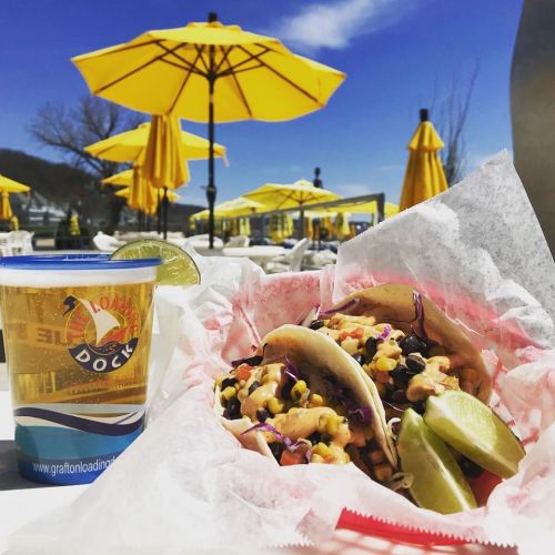 A plate of tacos with toppings and lime, alongside a drink, set against a sunny outdoor dining area with yellow umbrellas.