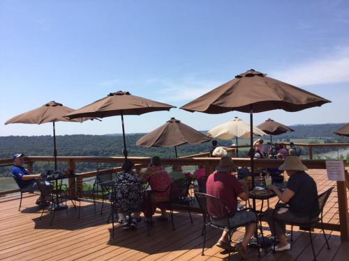 A sunny outdoor café with patrons sitting under umbrellas, overlooking a scenic view of hills and a river.
