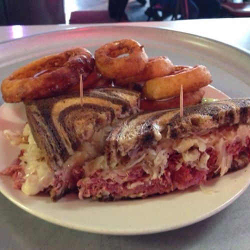 A plate with a stacked sandwich filled with corned beef and sauerkraut, accompanied by onion rings.