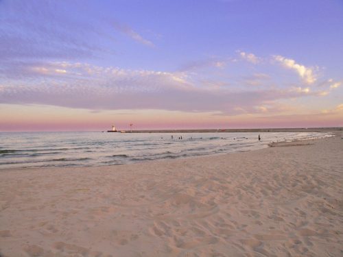 A serene beach at sunset, with gentle waves, soft sand, and a distant lighthouse under a colorful sky.