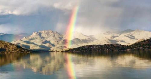 A vibrant rainbow arcs over mountains, reflecting in a calm lake under a cloudy sky.