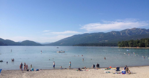 A sunny beach scene with people swimming and relaxing by a lake, surrounded by mountains and clear blue skies.