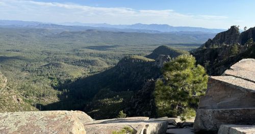 A panoramic view of lush green mountains and valleys under a clear blue sky, with rocky outcrops in the foreground.