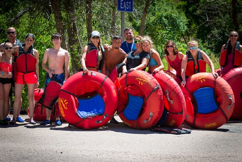 A group of people in swimsuits and life jackets pose with red inner tubes by a road sign in a sunny outdoor setting.