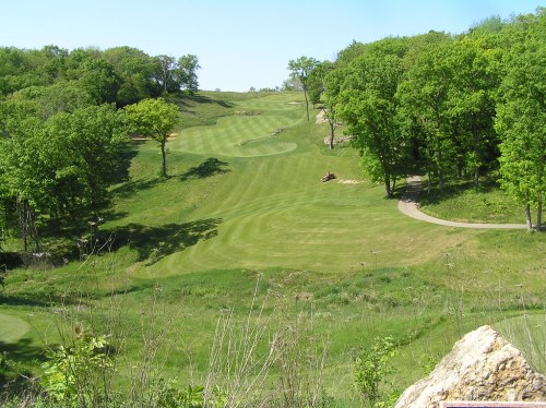 A scenic view of a golf course with rolling hills, lush green grass, and trees lining the fairways under a clear sky.