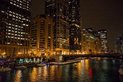 City skyline at night with illuminated buildings reflecting on a river, boats docked along the waterfront.