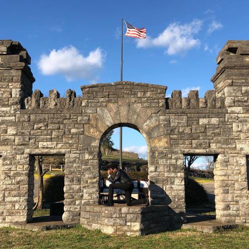 A person kneels in a stone archway of a castle-like structure, with an American flag waving above against a blue sky.