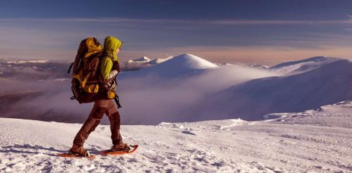A person snowshoeing on a snowy mountain landscape with a backpack, surrounded by peaks and clouds.