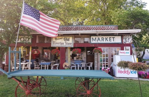 A quaint market with a vintage storefront, featuring a flag, flowers, and outdoor seating. Route 66 signage visible.