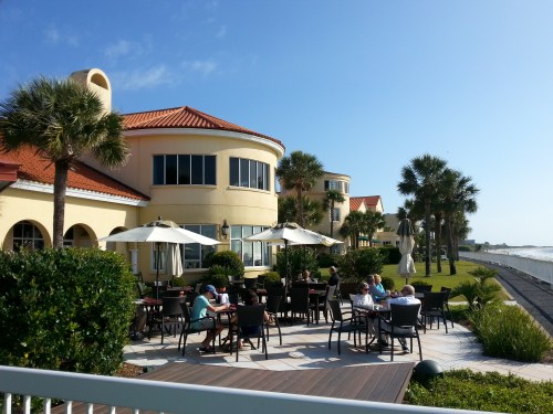 Outdoor dining area with palm trees and umbrellas, overlooking the ocean on a sunny day.