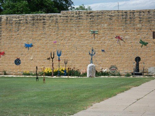 Colorful metal sculptures of dragonflies and garden tools adorn a stone wall, with flowers and grass in the foreground.