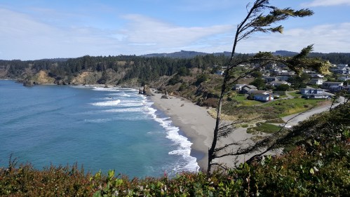 Coastal view featuring a sandy beach, waves, and a small town surrounded by lush greenery and hills.
