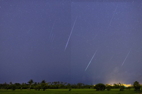 A night sky filled with stars and multiple shooting stars over a dark landscape with distant lights.