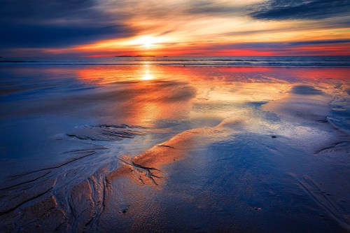 A vibrant sunset reflects on wet sand, creating a colorful blend of orange, blue, and purple hues over the ocean.