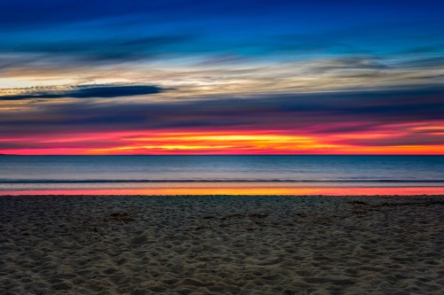 A vibrant sunset over the ocean, with colorful clouds reflecting on the water and sandy beach in the foreground.