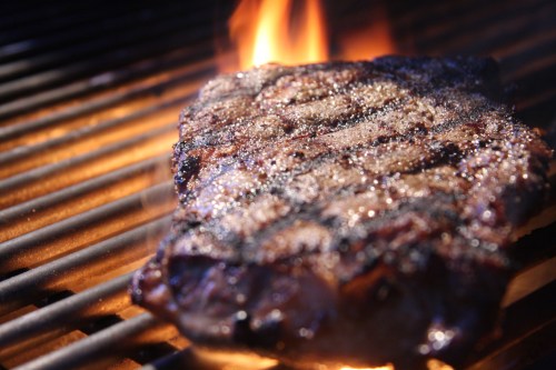 A juicy steak sizzling on a grill, with flames licking the edges and grill marks visible on the surface.