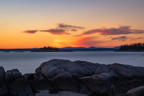 A serene coastal scene at sunset, featuring rocky shores, calm waters, and distant mountains under a colorful sky.