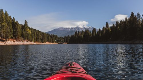 A red kayak on calm water, surrounded by tall trees and mountains under a clear blue sky.