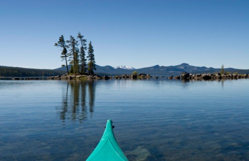 A kayak's bow points towards a calm lake with trees and mountains reflected in the water under a clear blue sky.