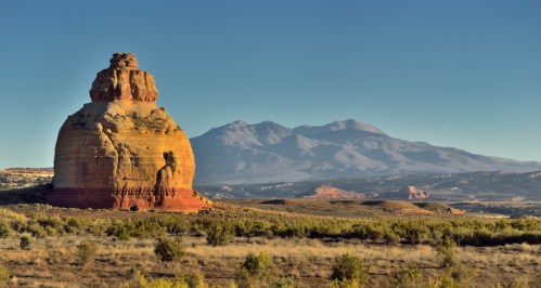 A large, rounded rock formation stands in the foreground with mountains in the background under a clear blue sky.