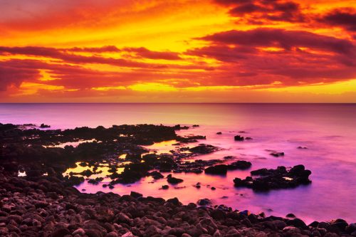 A vibrant sunset over a rocky shoreline, with colorful clouds reflecting on the calm water.