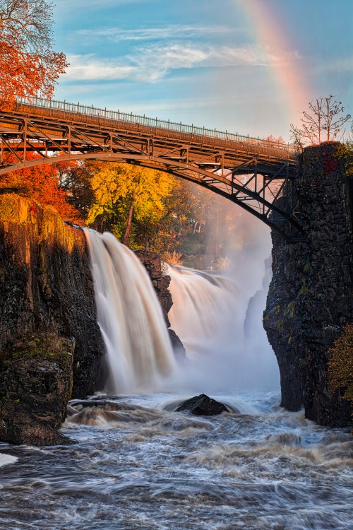 A scenic view of a waterfall under a bridge, surrounded by autumn foliage and a rainbow in the sky.