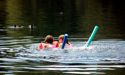 Two children swim in a lake, using pool noodles, with ducks floating nearby.