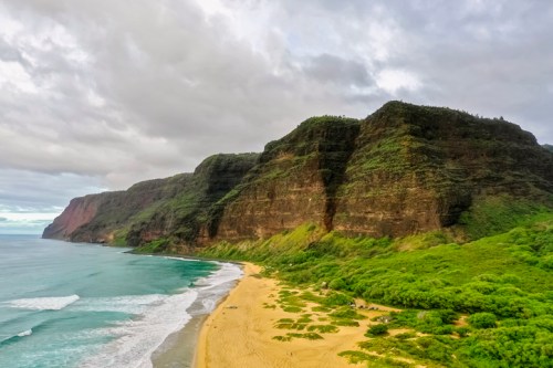 A scenic view of a sandy beach bordered by lush green cliffs under a cloudy sky. Waves gently crash on the shore.