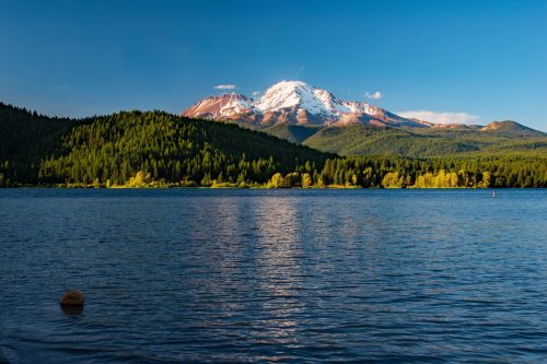 A serene lake reflects a snow-capped mountain surrounded by lush green forests under a clear blue sky.