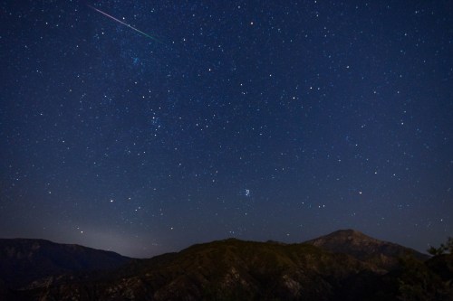 A starry night sky over mountains, featuring a shooting star and clusters of stars scattered across the dark blue expanse.