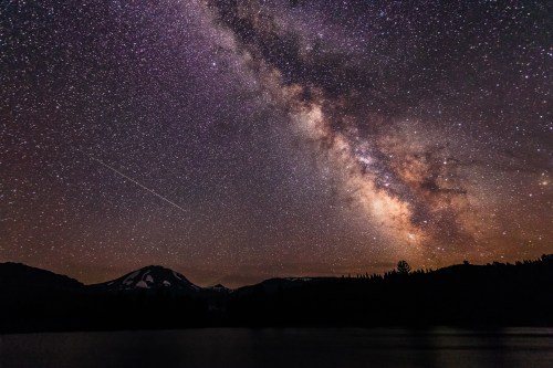 A stunning night sky filled with stars and the Milky Way, above a mountain landscape and a calm lake.